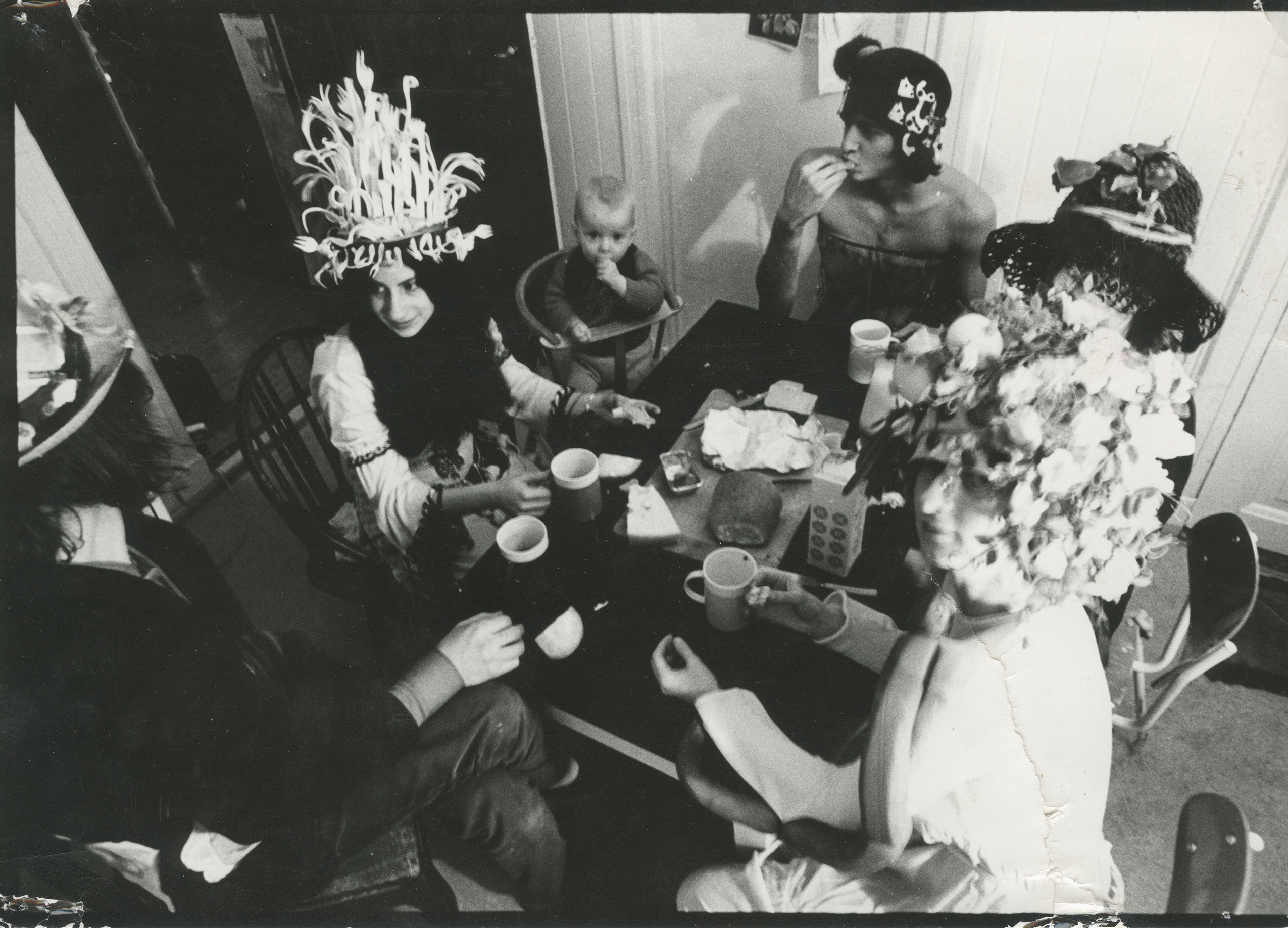 A black-and-white photograph of a group of people sitting around a dining table, drinking from mugs and sharing food. Several adults wear elaborate and whimsical hats decorated with flowers, paper, or objects, while a baby sits in a high chair at the table.