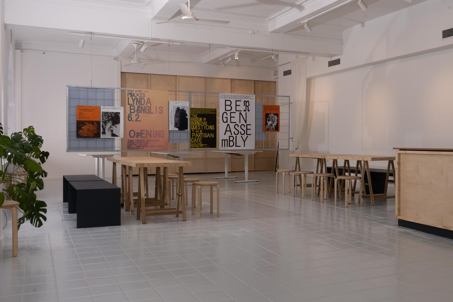 Minimalist gallery space with white walls, high ceilings, and large arched windows. Wooden tables with matching stools are placed in the center, alongside a few black benches. Posters with bold typography and graphic designs are displayed on wire grids against the back wall, promoting art events and exhibitions. A green potted plant sits near the window, adding a touch of color to the clean, modern interior.