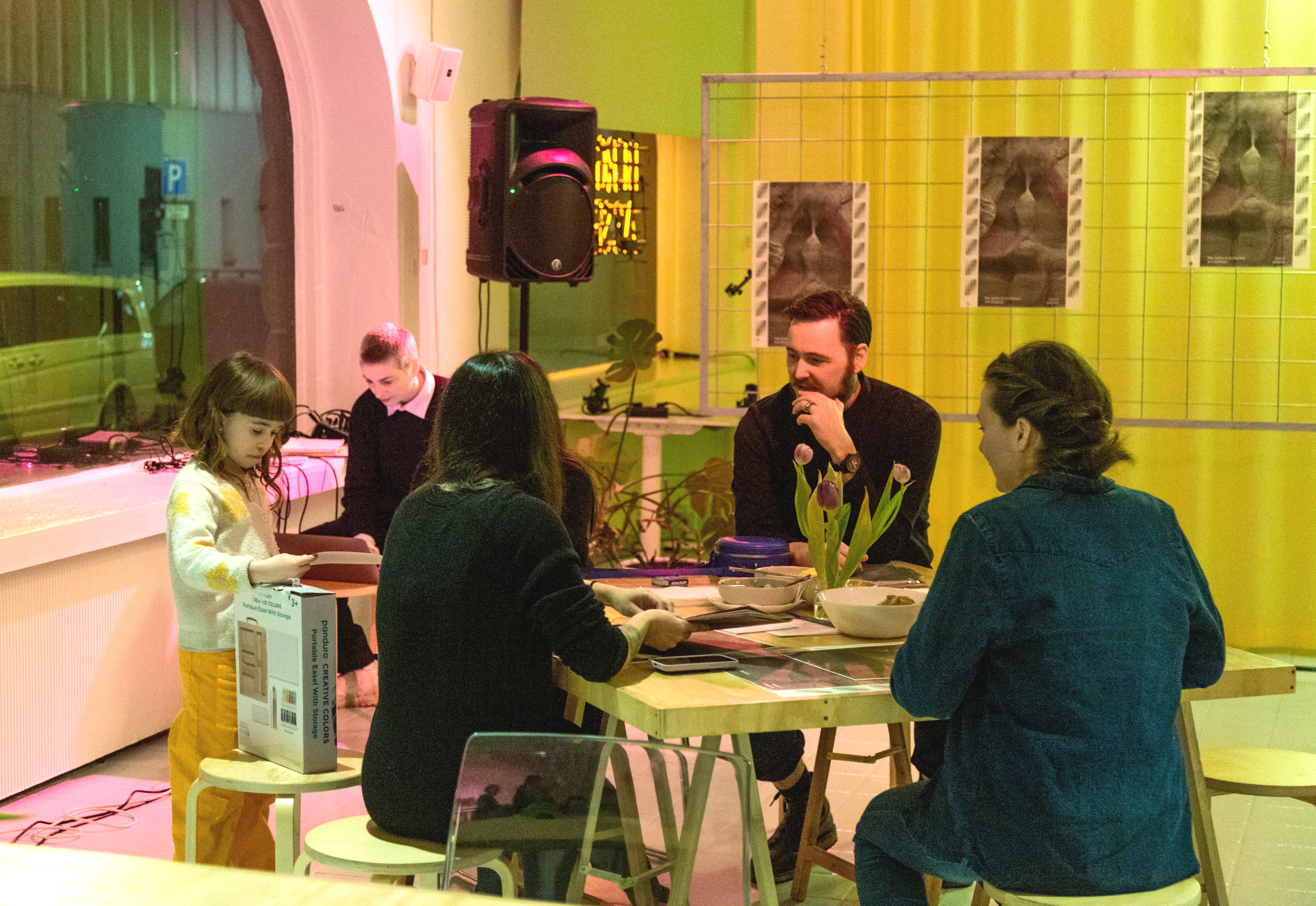 People gathered around a table in a brightly lit room, engaging in a creative or social activity; a child stands nearby holding a box.