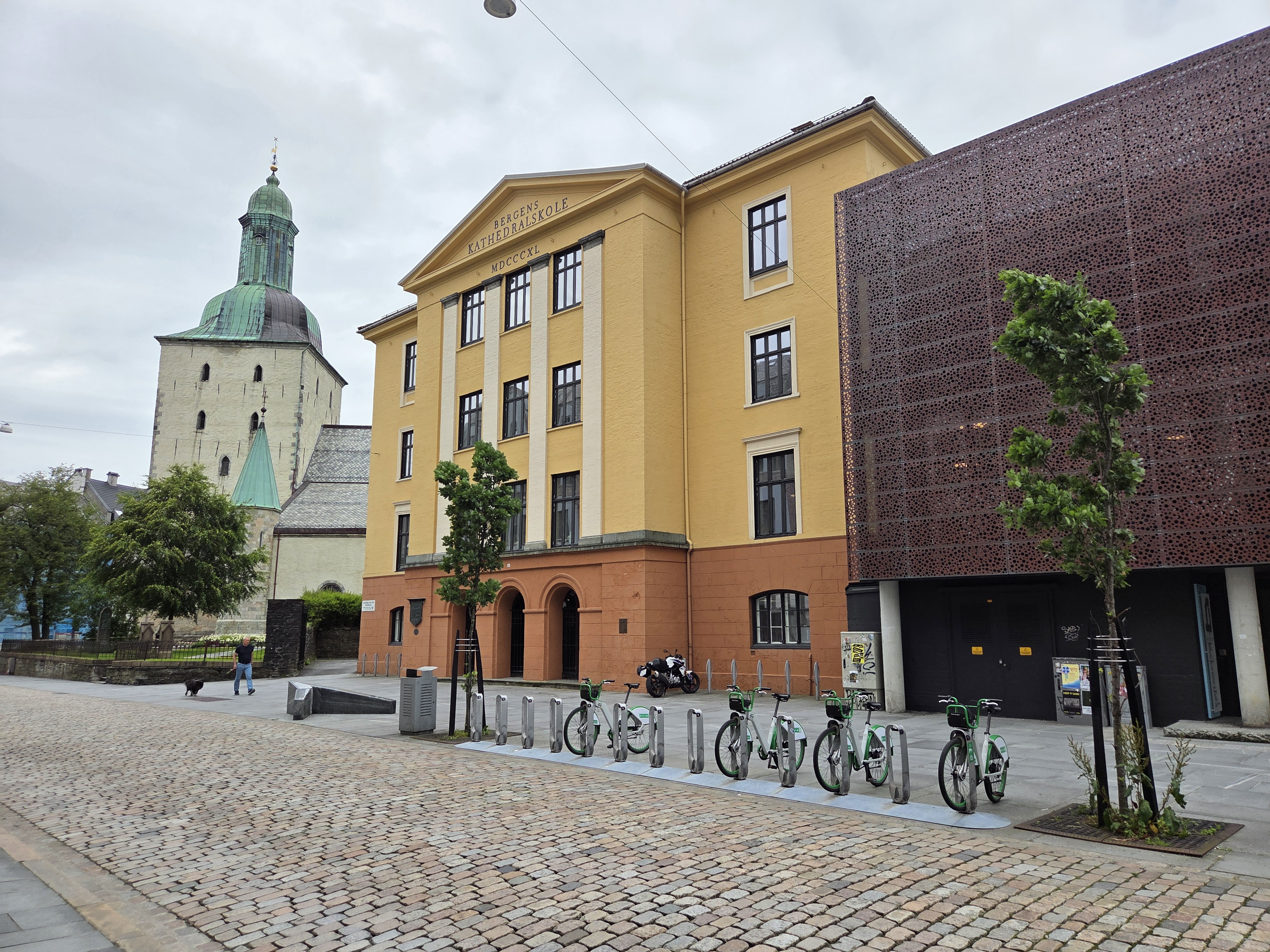 Bergen Cathedral School, a historic yellow and orange building with arched entrances and tall windows, stands next to a modern structure with a perforated metal facade.