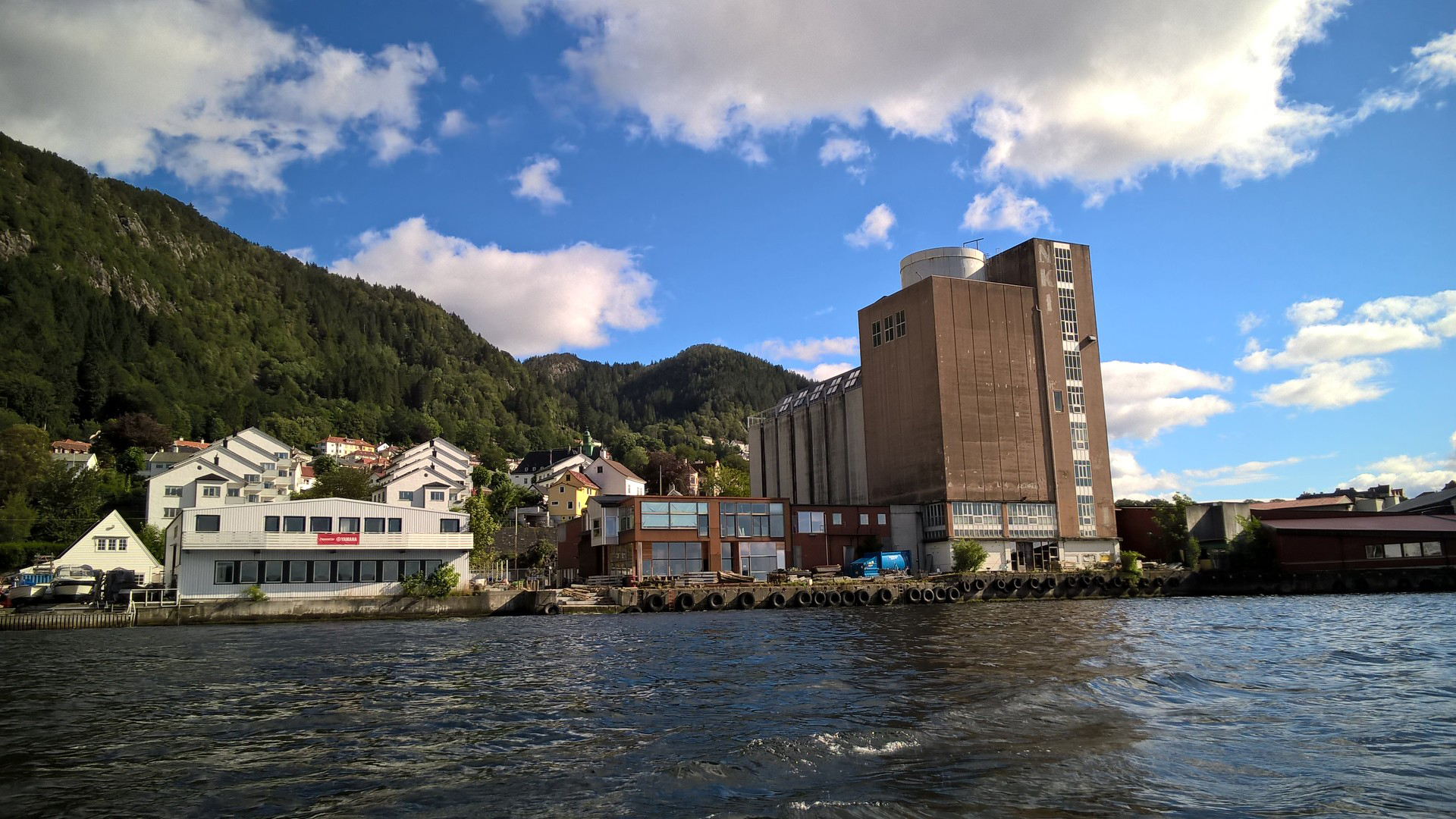 A large, tall industrial-style building with a tower and adjoining structures, located by the waterfront with forested hills in the background.