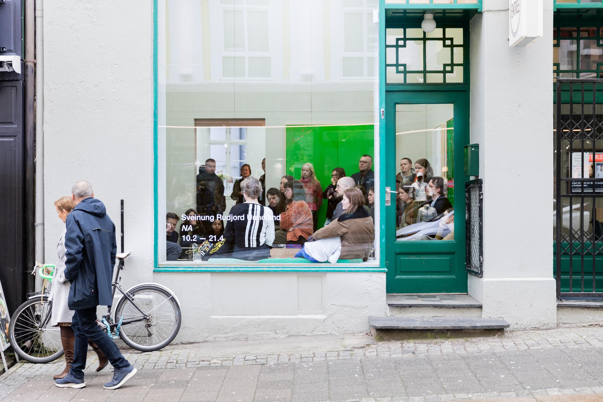 People gathered inside a gallery space, seen through a large front window with green trim.