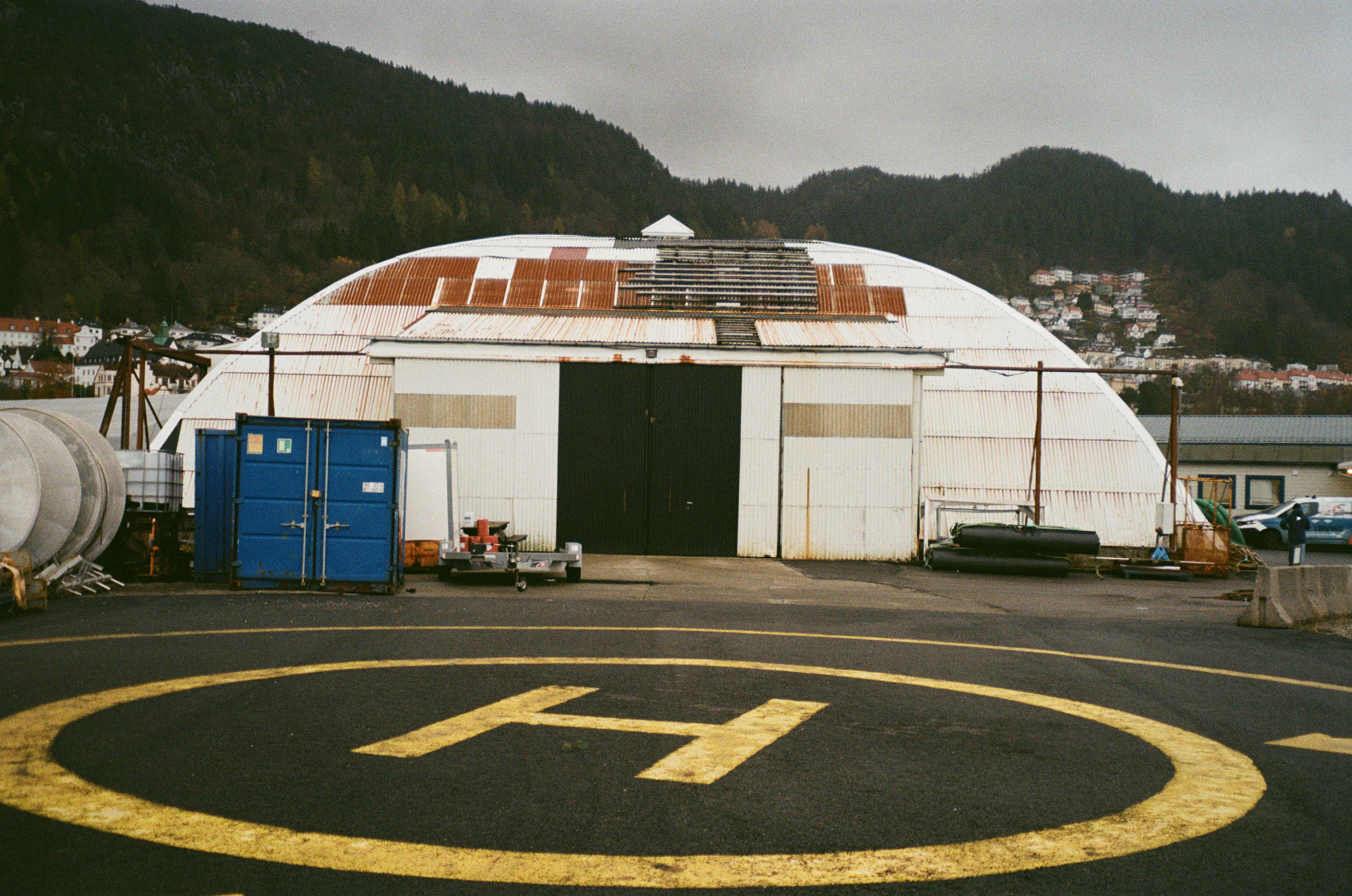 Old hangar with rusted roof behind a helipad marked with a yellow “H.”