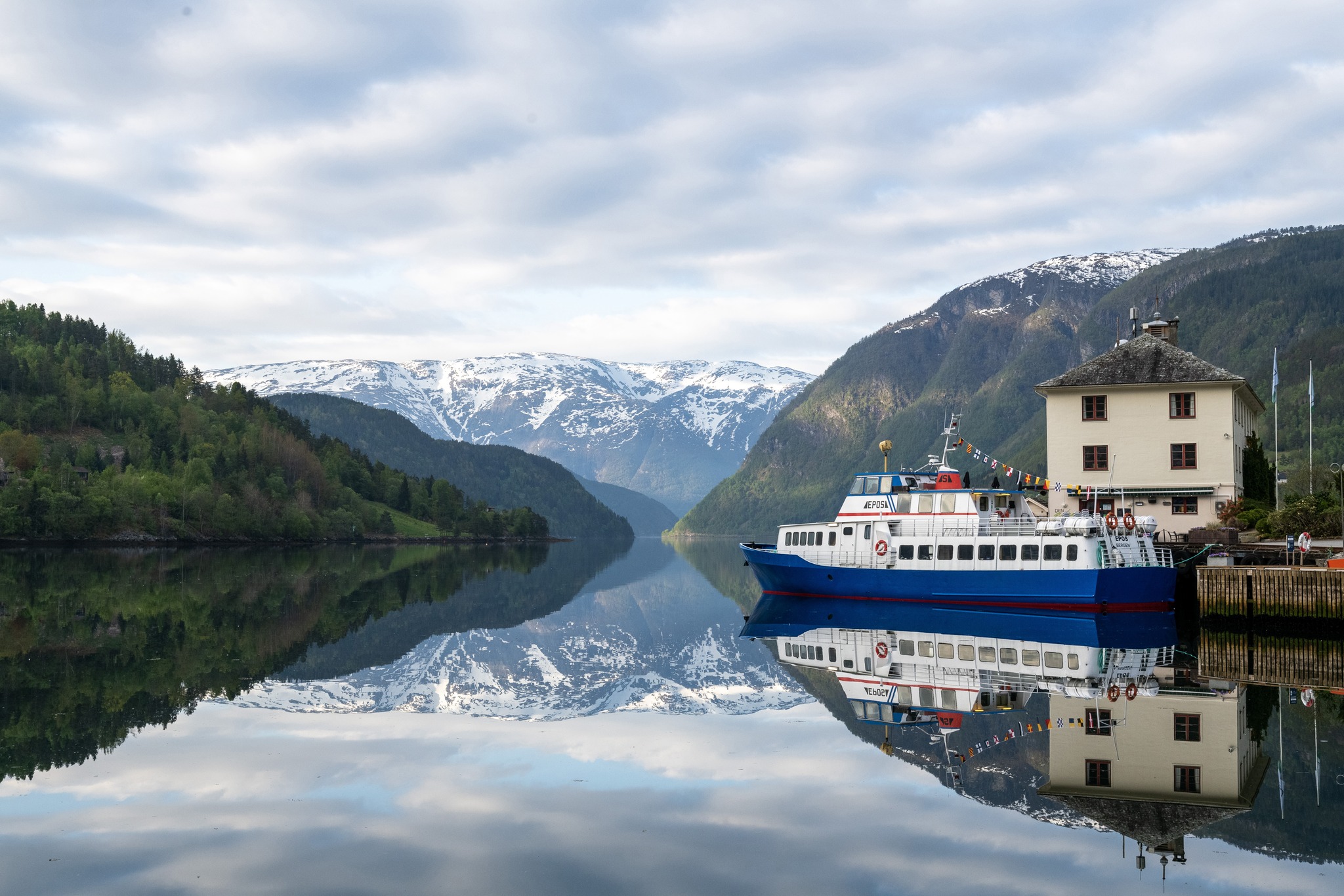A white and blue passenger boat docked by a building on a calm fjord.