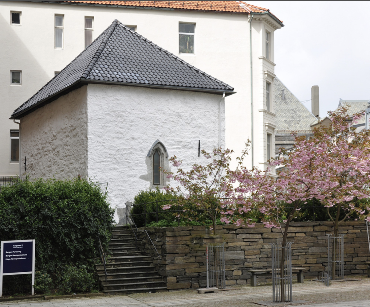 Small white stone building with a dark tiled roof, beside blooming pink trees.