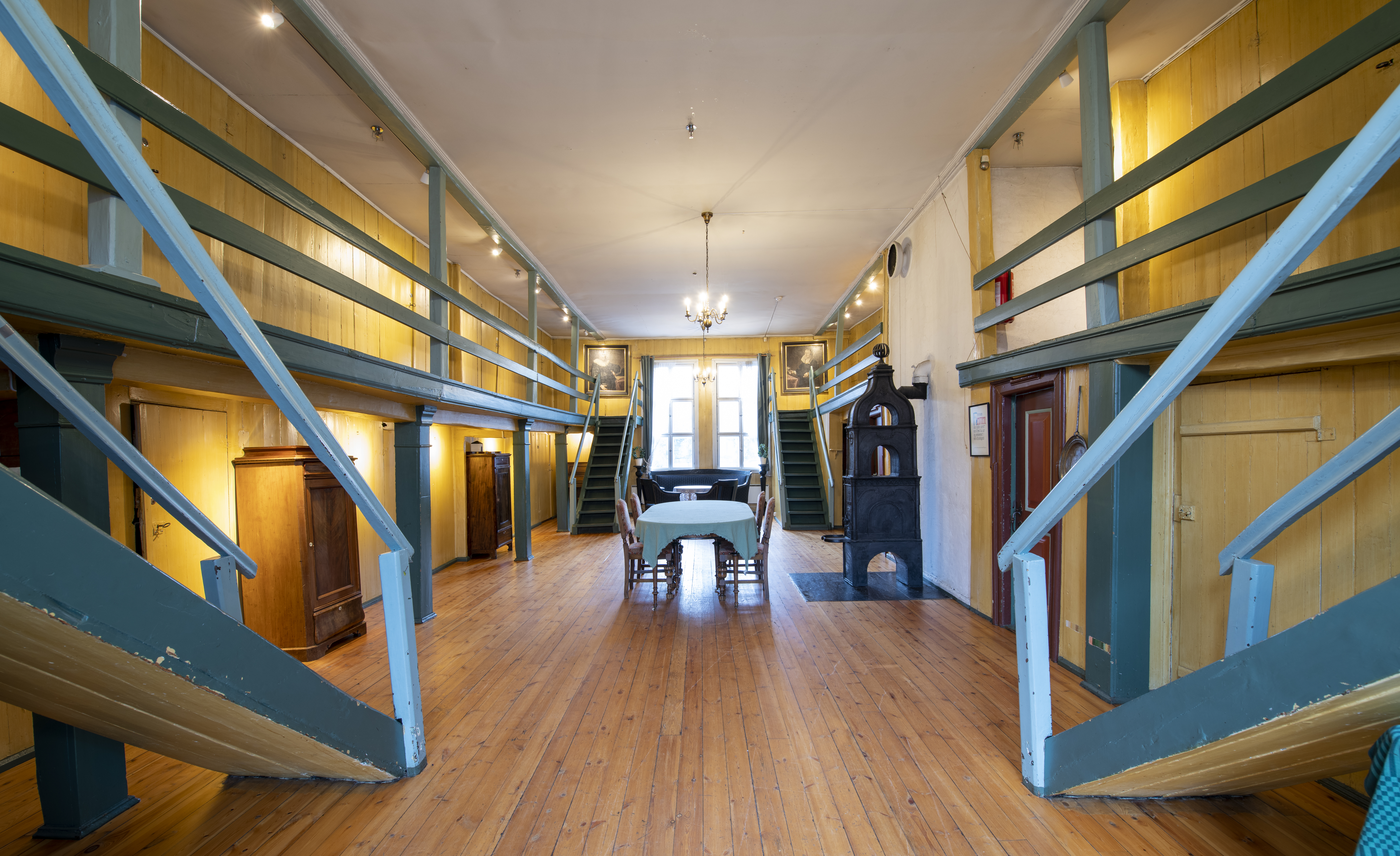 Historic wooden interior with long tables, staircases on both sides, and a black cast-iron stove