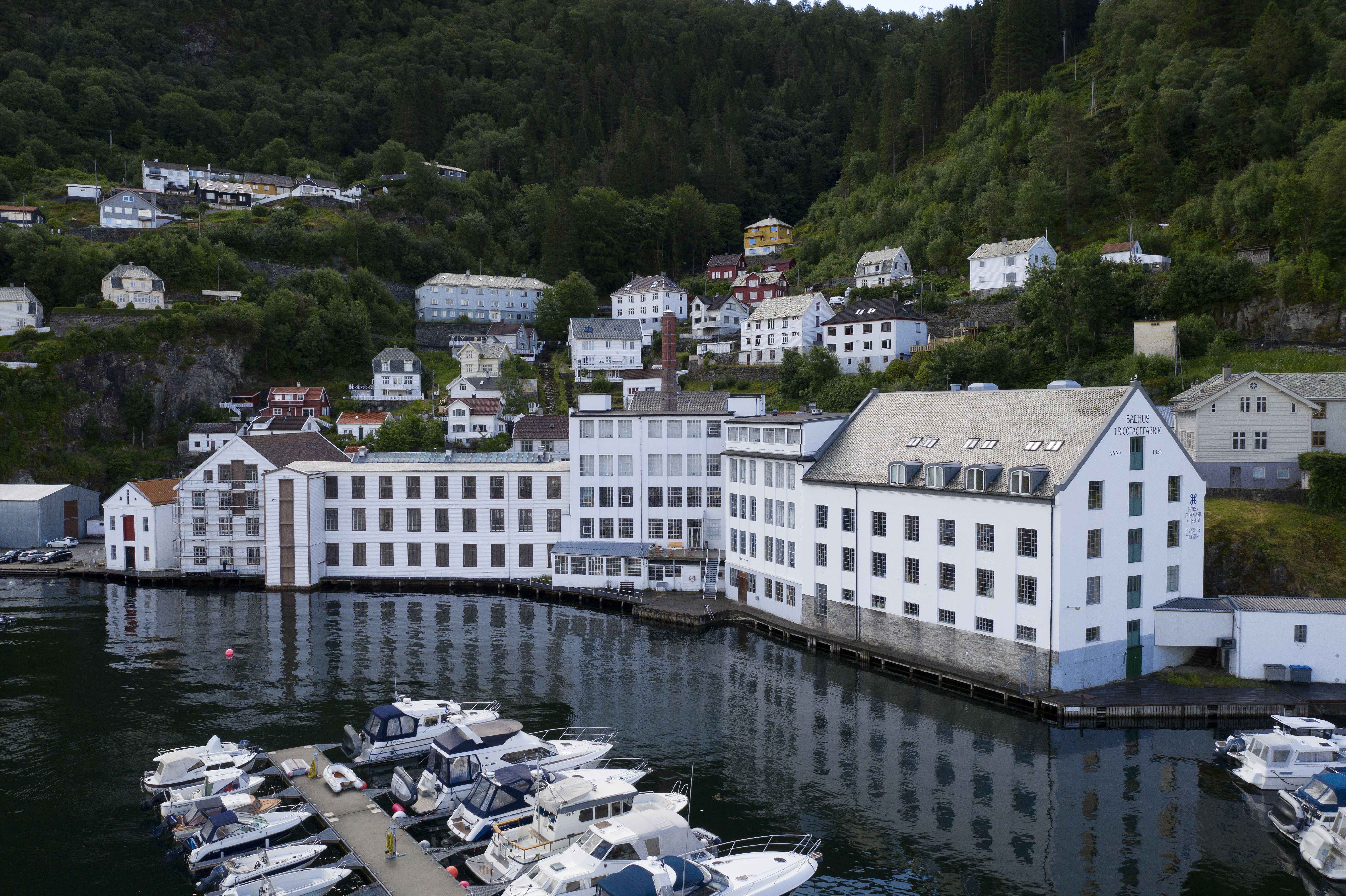 Large white former factory building by the waterfront, with small boats docked in front and houses on the hillside behind.